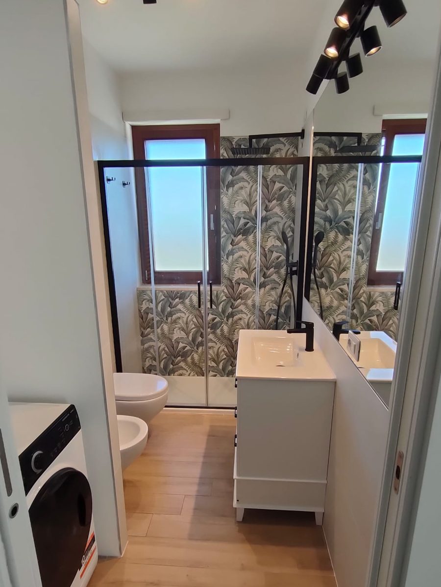 Dopo: lo stesso bagno con doccia walk-in, parete a foglie tropicali e mobile bianco